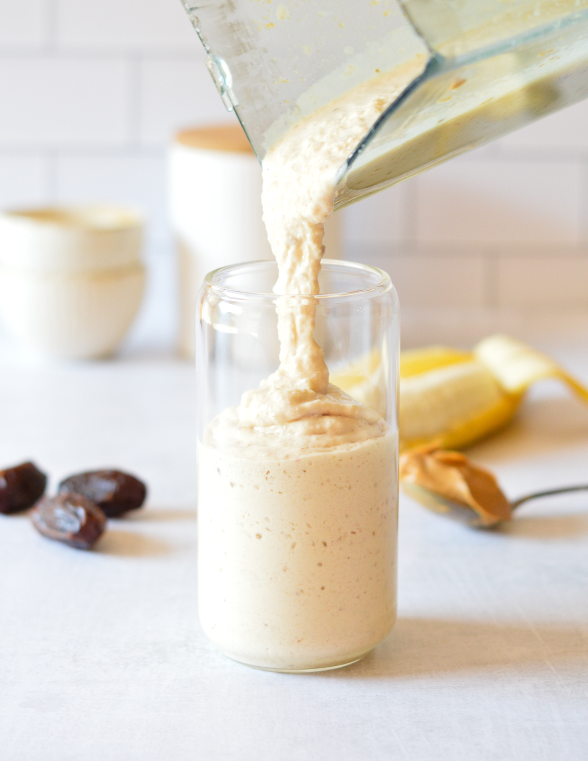 pouring a peanut butter smoothie into a glass next to dates, banana, and peanut butter.